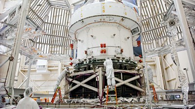 Two technicians in protective suits work on an Orion spacecraft from NASA. The spacecraft is situated in a large clean room with scaffolding and equipment surrounding it. The technicians appear to be inspecting or assembling various components.