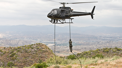 An Airbus Helicopter hovers above a rugged, mountainous landscape. Two ropes extend from the helicopter, with a person descending on one of them. The background shows a mix of vegetation and dry terrain with mountains in the distance under a cloudy sky.