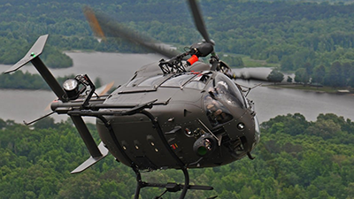 A military helicopter flies over a lush, forested landscape with a river visible below. The U.S. Army awards Airbus contract for helicopter modernization, showcasing advanced equipment attached and rotors in motion as it hovers in mid-air, slightly tilted.