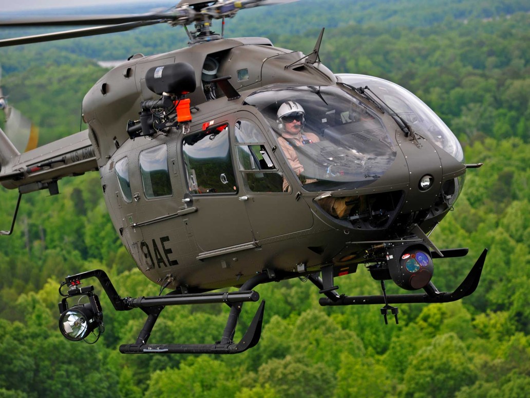 A military helicopter with a pilot in the cockpit hovers in the air above a dense green forest. The helicopter, equipped with advanced surveillance and navigation equipment on its exterior, appears to be in mid-flight. The sky is clear, and visibility is good.