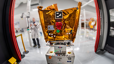 A worker in a lab coat stands next to a cube-shaped scientific instrument covered in gold foil, mounted on a wheeled platform, inside a clean room with red-framed entryway. The backdrop of laboratory equipment and another worker in white attire reflects the high-tech environment where Airbus announces its purchase of Eutelsat OneWeb's stake.