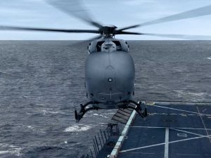 A gray UH-72 helicopter is seen in mid-air, just above the deck of a ship over a vast expanse of ocean. The helicopter's blades are spinning, and the ship's deck shows markings and structures typical of a naval vessel. The sky is cloudy with a calm sea.