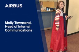 A woman in traditional attire stands smiling near a door, identified as Molly Townsend, Head of Internal Communications, with the Airbus logo displayed above her name. Celebrating Asian-American Pacific Islander Heritage Month, she wears a red and white traditional dress. Office furniture is visible in the background.