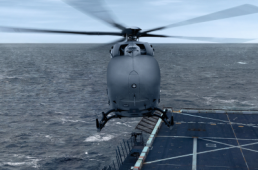A helicopter hovers over the edge of an aircraft carrier at sea. The gray helicopter, possibly part of a USMC Aerial Logistics Connector contract, has its rotors in motion, while the vast ocean extends to the horizon under a cloudy sky. The aircraft carrier's deck is partially visible with markings and a safety net.